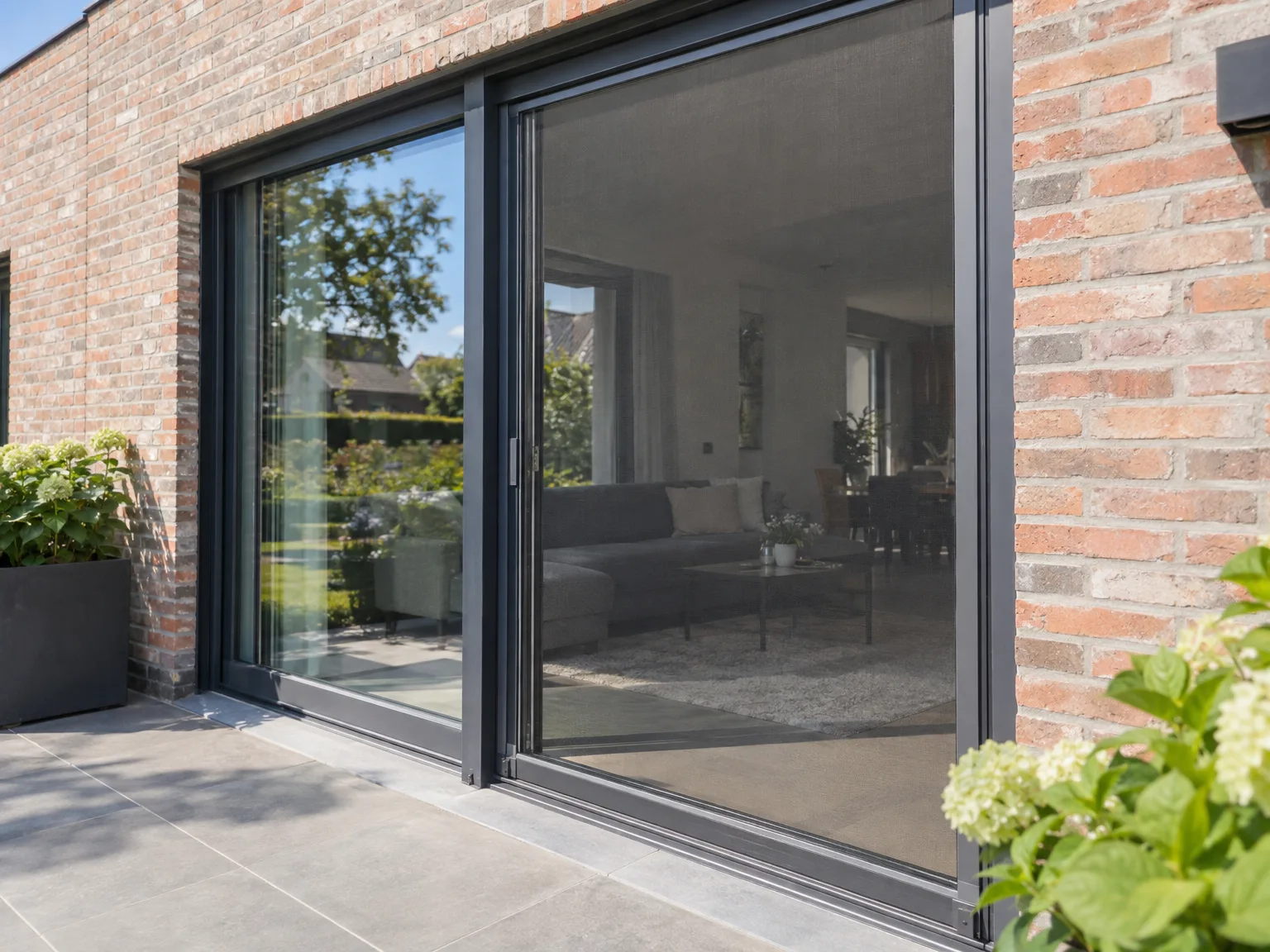 Grote aluminium schuiframen in donkergrijs geopend op terras tegen bakstenen woninggevels